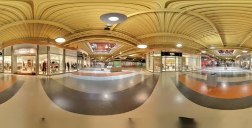 A panoramic view of a modern shopping mall with various storefronts and a spacious, well-lit interior.