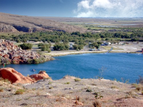 A scenic view of a blue lake surrounded by rocky terrain and greenery, with a distant parking area and hills in the background.