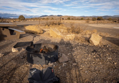 A campfire burns in a fire pit surrounded by rocky terrain and sparse vegetation under a blue sky.