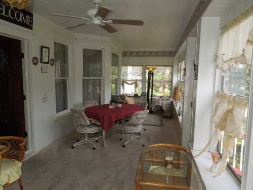 A cozy sunroom with a round table, chairs, and large windows, decorated with curtains and a welcoming sign.