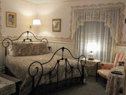 Cozy bedroom with a wrought iron bed, lace curtains, floral decor, and a vintage armchair by a lamp-lit nightstand.