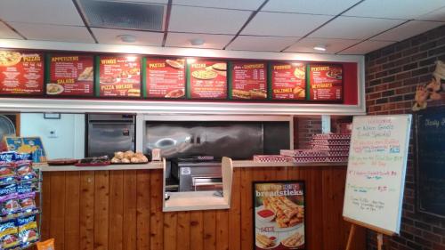 Menu board displaying various food items at a restaurant, with a counter and pizza boxes in the background.