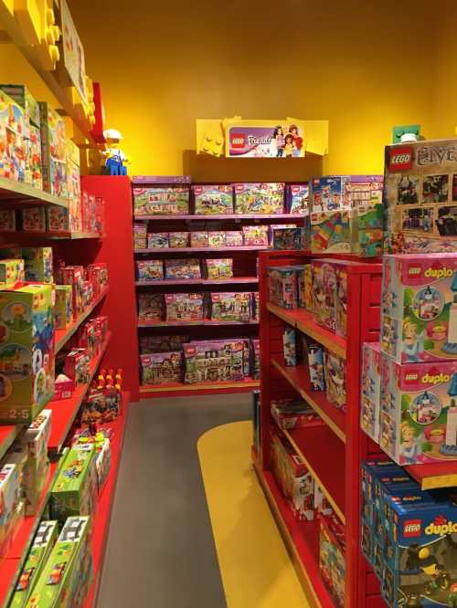 A colorful LEGO store aisle filled with various LEGO sets on red shelves against a bright yellow wall.