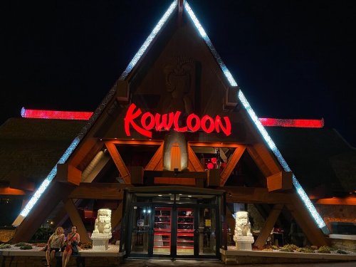 A brightly lit restaurant entrance with a triangular roof, featuring the name "Kowloon" in red neon lights at night.