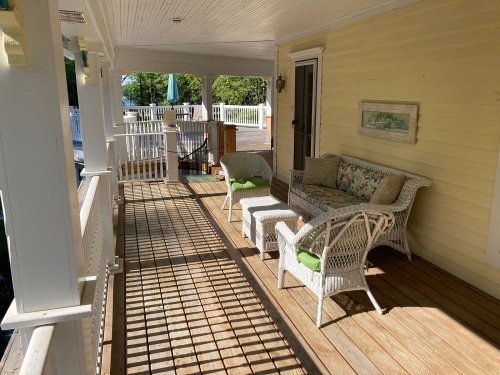 A sunny porch with wicker furniture, including a sofa and chairs, surrounded by white railings and wooden flooring.