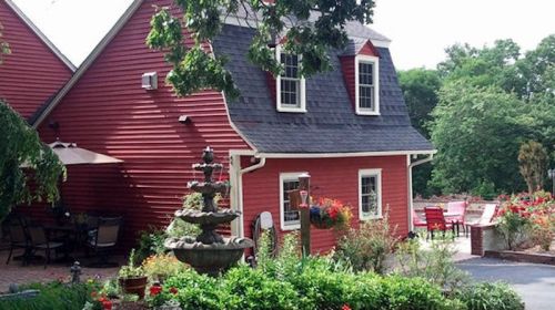 A charming red house with a sloped roof, surrounded by greenery and colorful flowers, featuring a decorative fountain.