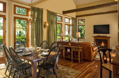 Cozy dining area with a wooden table set for a meal, adjacent to a living room with a fireplace and large windows.