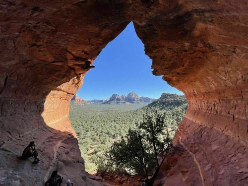 View through a rock formation, framing a landscape of mountains and trees under a clear blue sky.