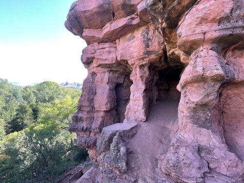 Rocky cliff formation with pinkish hues, featuring a cave entrance and surrounding greenery.
