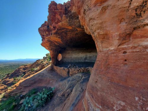 A rocky cave with a stone wall, overlooking a vast desert landscape under a clear blue sky.