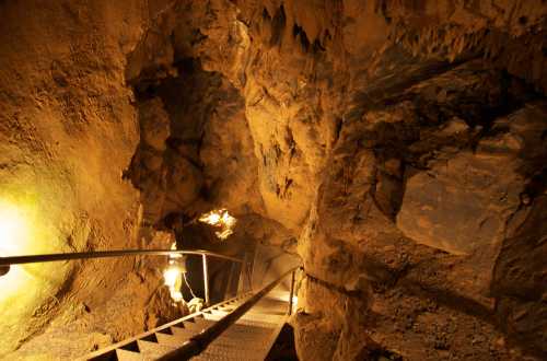 A dimly lit cave with a metal staircase leading down into rocky terrain. Stalactites and rough walls are visible.