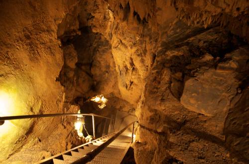 A dimly lit cave with a metal staircase leading down into rocky terrain. Stalactites and rough walls are visible.