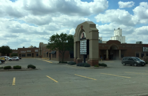 A vacant shopping plaza with a sign listing businesses, surrounded by a parking lot and cloudy sky.