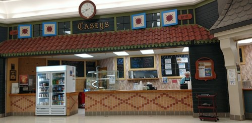 A food counter named "Casey's" in a mall, featuring a menu board, display case, and a clock above.