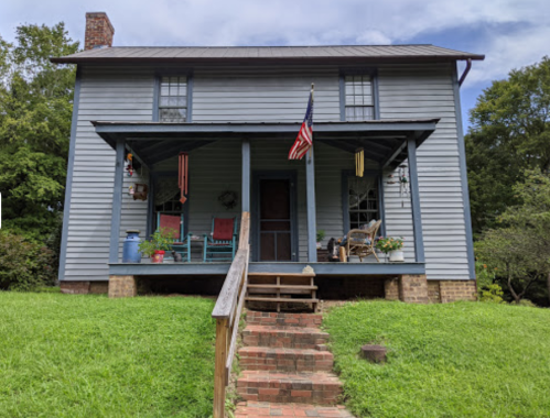 A blue two-story house with a porch, American flag, and colorful chairs, surrounded by greenery and a brick walkway.