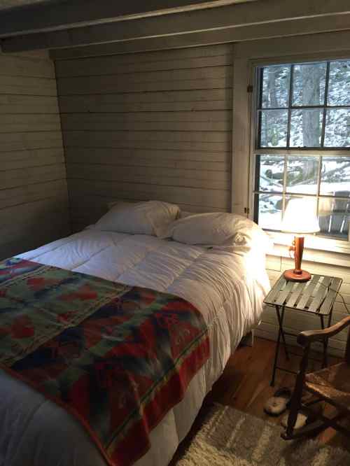 Cozy bedroom with a bed, colorful blanket, wooden chair, and a lamp by a window showing a snowy landscape outside.