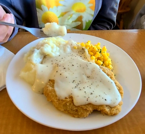 A plate of fried chicken smothered in gravy, served with mashed potatoes and corn. A fork is poised to take a bite.