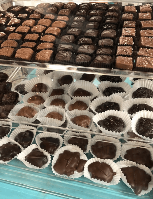 A display of assorted chocolates in a glass case, featuring various shapes and coatings, arranged in paper liners.