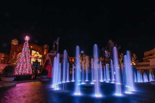 A festive scene at night featuring a lit Christmas tree and colorful fountains in front of a decorated building.