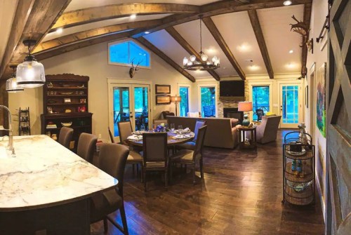 Cozy open-concept living space with wooden beams, a dining area, and a fireplace, featuring large windows and warm decor.