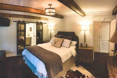 Cozy bedroom with a large bed, wooden beams, lamps, and a rustic decor style. A mirror and dresser are visible.