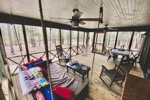 Cozy screened porch with rocking chairs, a swing, and colorful cushions, surrounded by trees and nature.