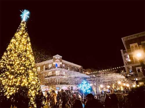 A festive night scene with a brightly lit Christmas tree and snowflakes falling over a bustling holiday market.