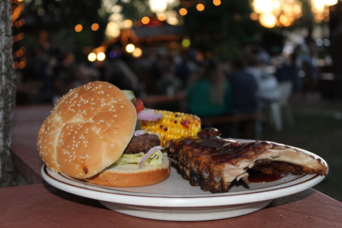 A plate with a hamburger and ribs, accompanied by corn, set against a blurred outdoor gathering at sunset.