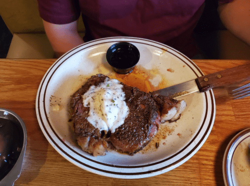 A plate with a seasoned steak topped with creamy sauce, accompanied by a small cup of dipping sauce.