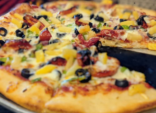 A close-up of a pizza topped with pepperoni, olives, pineapple, and green peppers, with a slice being lifted.