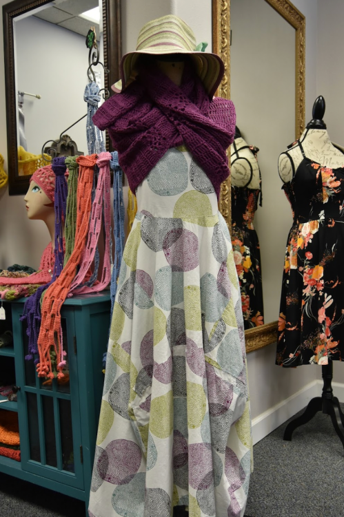 A mannequin displays a patterned dress with a purple shawl, surrounded by colorful accessories and a mirror.