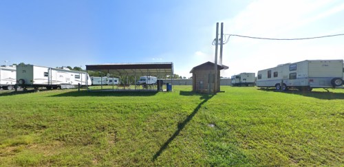 A grassy area with several RVs parked, a covered shelter, and a small utility building under a clear blue sky.