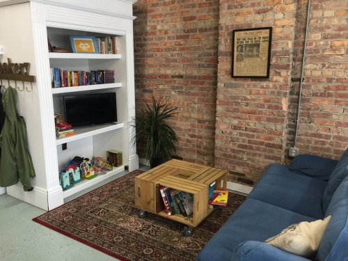 Cozy living space with a blue couch, wooden coffee table, bookshelf, TV, and a plant against a brick wall.