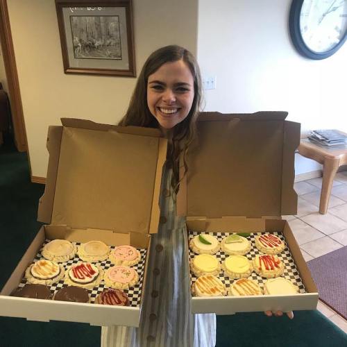 A smiling woman holds two large boxes of colorful cupcakes, showcasing a variety of flavors.