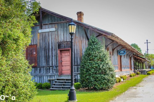 A rustic wooden building with a red door, surrounded by greenery and a lamp post, set against a clear sky.