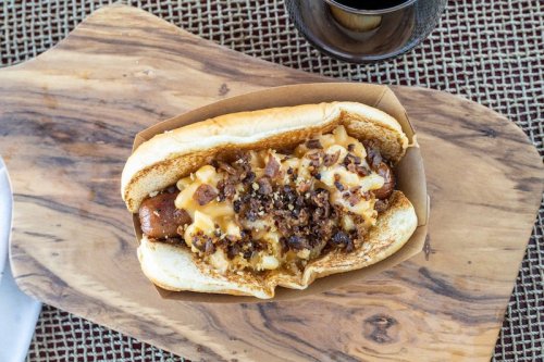 A hot dog in a bun topped with cheese and crumbled bacon, served on a wooden board with a drink in the background.