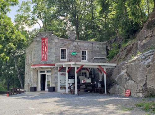 A stone building with a red "Open" sign, surrounded by trees and rocky terrain, featuring outdoor seating.