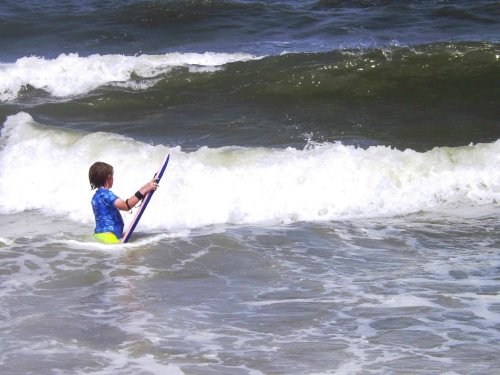 A child in a blue shirt and yellow shorts stands in the ocean, holding a boogie board as waves crash nearby.