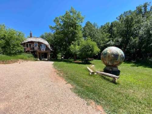 A rustic house with a round stone sculpture in a green, wooded area under a clear blue sky.