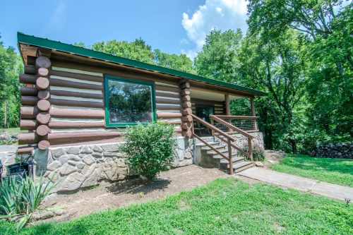 A log cabin with a stone foundation, surrounded by greenery and trees, featuring a front porch and large window.