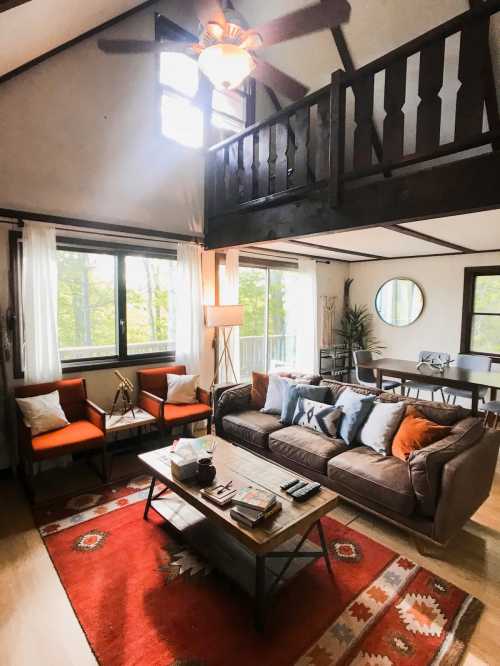 Cozy living room with a brown sofa, orange chairs, a coffee table, and large windows overlooking greenery.