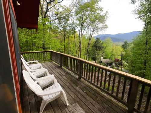 A wooden deck with white chairs overlooks a lush green landscape and distant mountains under a cloudy sky.