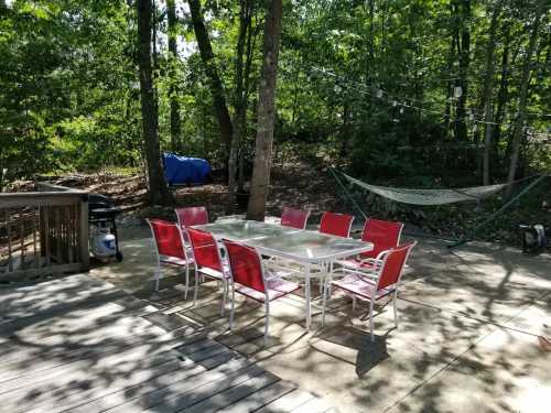 A patio with a large table surrounded by red chairs, set in a wooded area with a hammock in the background.