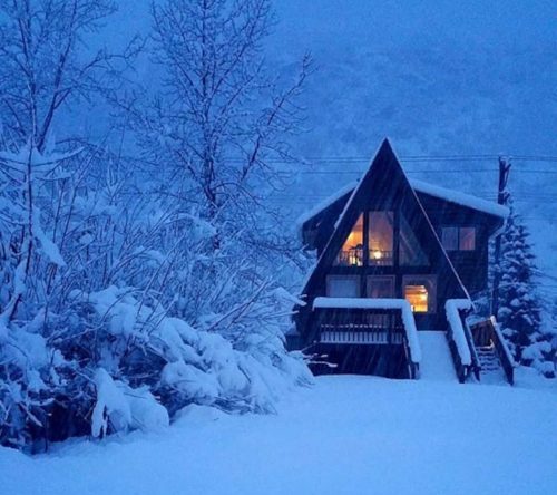 A cozy A-frame house surrounded by deep snow, with warm light glowing from the windows in a winter landscape.