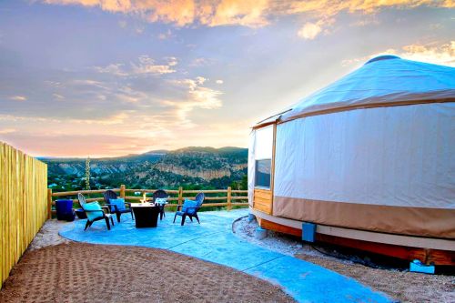 A cozy yurt with outdoor seating and a fire pit, set against a scenic mountain sunset.