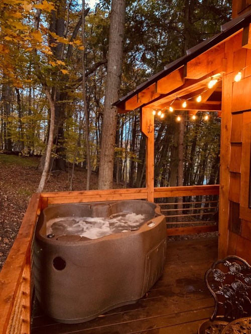 A cozy hot tub on a wooden deck surrounded by trees, illuminated by string lights in a serene autumn setting.