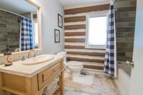 A modern bathroom featuring a wooden vanity, striped shower curtain, and log-style accent wall.