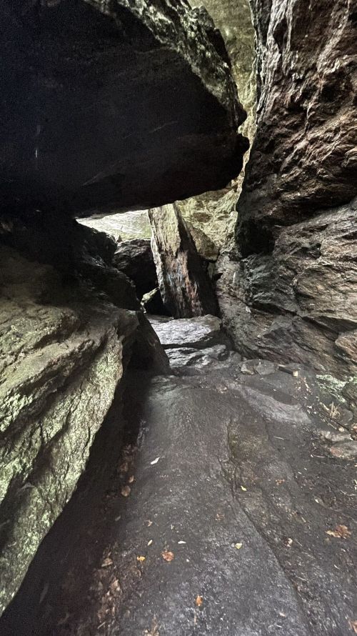 A narrow passage between large, dark rocks in a cave, with a rocky floor and faint light illuminating the entrance.