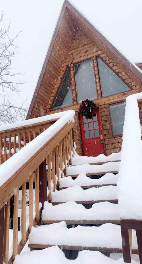A snowy scene featuring a wooden A-frame house with a red door and a wreath, surrounded by falling snow.