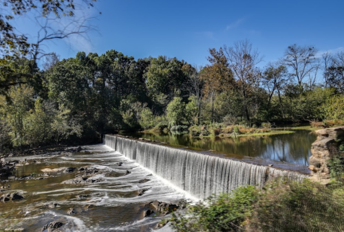 A serene waterfall cascades over a stone dam, surrounded by lush greenery and trees under a clear blue sky.